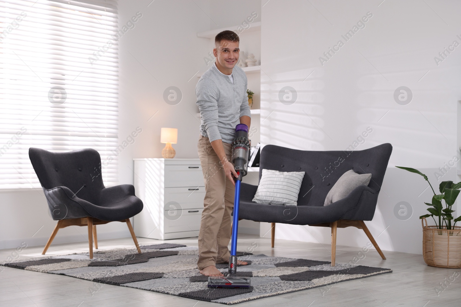 Photo of Young man cleaning rug with cordless vacuum cleaner at home