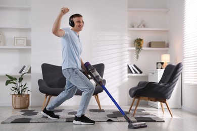 Photo of Young man in headphones having fun while cleaning floor with cordless vacuum cleaner at home