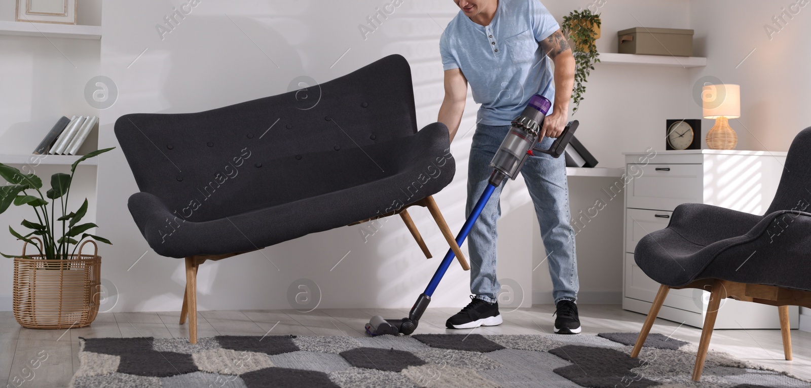 Photo of Young man cleaning rug under sofa with cordless vacuum cleaner at home, closeup