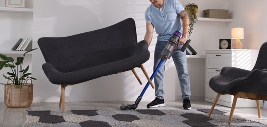 Photo of Young man cleaning rug under sofa with cordless vacuum cleaner at home, closeup