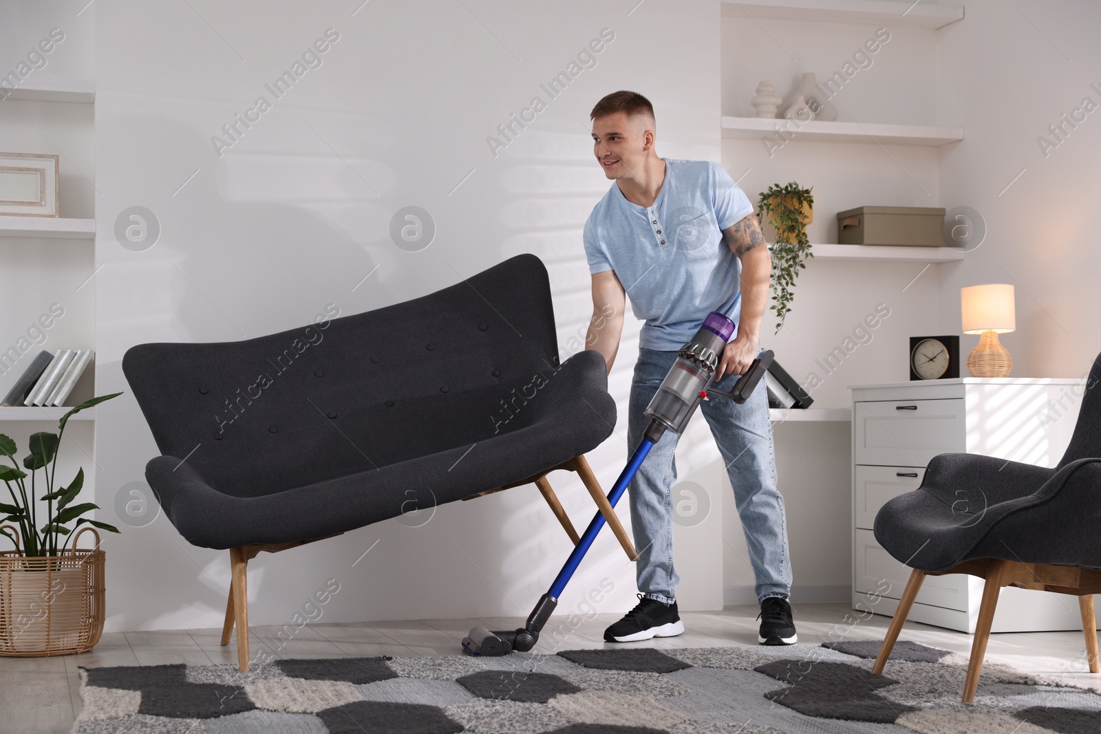 Photo of Young man cleaning rug under sofa with cordless vacuum cleaner at home