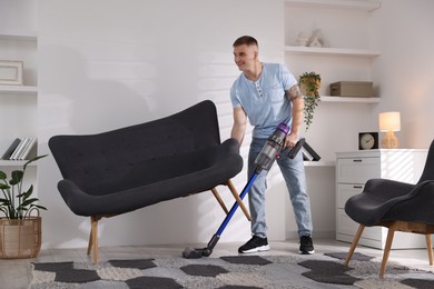 Photo of Young man cleaning rug under sofa with cordless vacuum cleaner at home