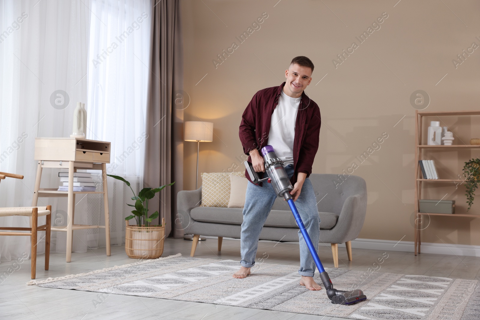 Photo of Young man cleaning rug with cordless vacuum cleaner at home