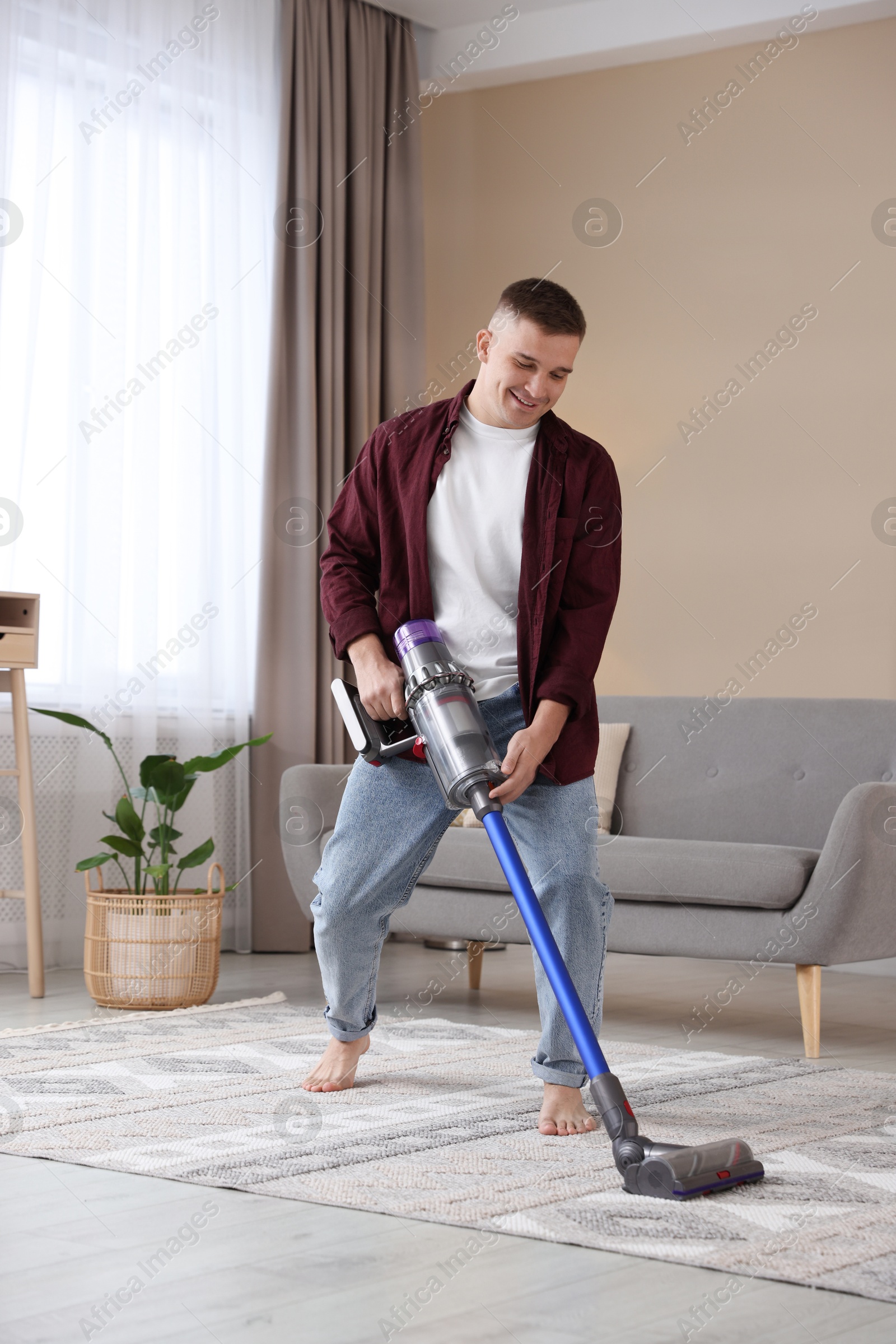 Photo of Young man cleaning rug with cordless vacuum cleaner at home