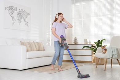 Photo of Teenage girl in headphones cleaning floor with cordless vacuum cleaner and her cute Chihuahua dog sitting in armchair at home