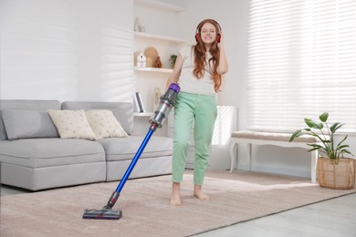 Photo of Teenage girl in headphones cleaning rug with cordless vacuum cleaner at home