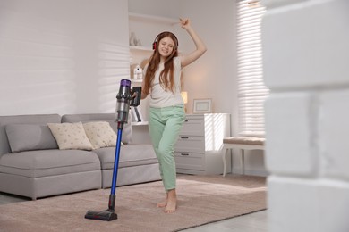 Photo of Teenage girl in headphones cleaning rug with cordless vacuum cleaner at home