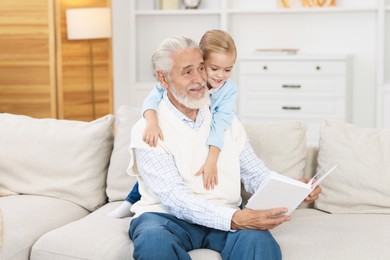 Grandpa and his granddaughter reading book together on sofa at home