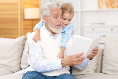 Photo of Grandpa and his granddaughter reading book together on sofa at home