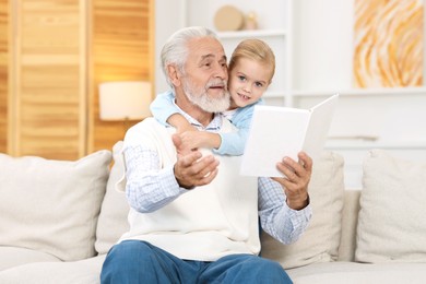 Photo of Grandpa and his granddaughter reading book together on sofa at home