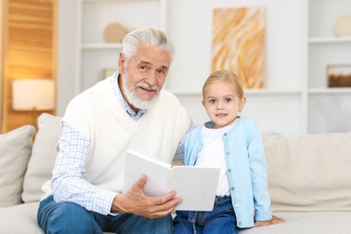 Photo of Grandpa and his granddaughter reading book together on sofa at home