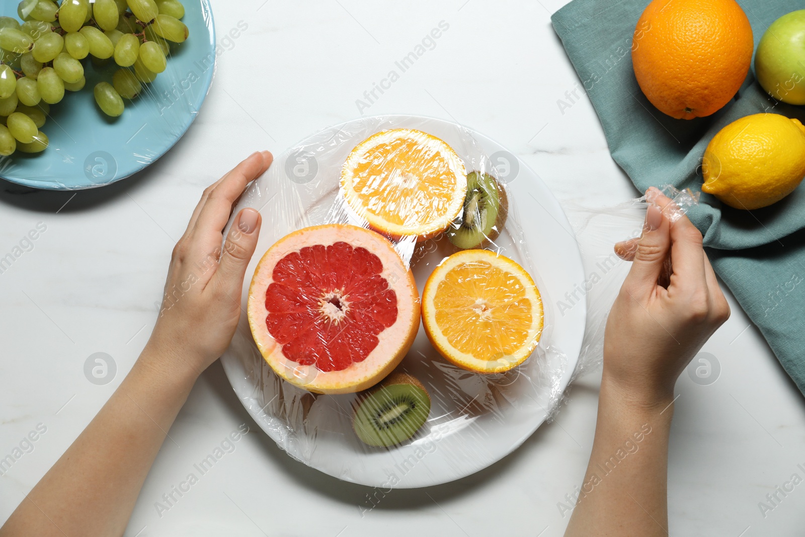 Photo of Woman putting plastic food wrap over plate with fresh fruits at white marble table, top view