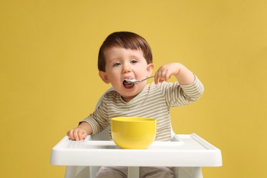 Photo of Cute little kid eating healthy baby food from bowl in high chair on yellow background