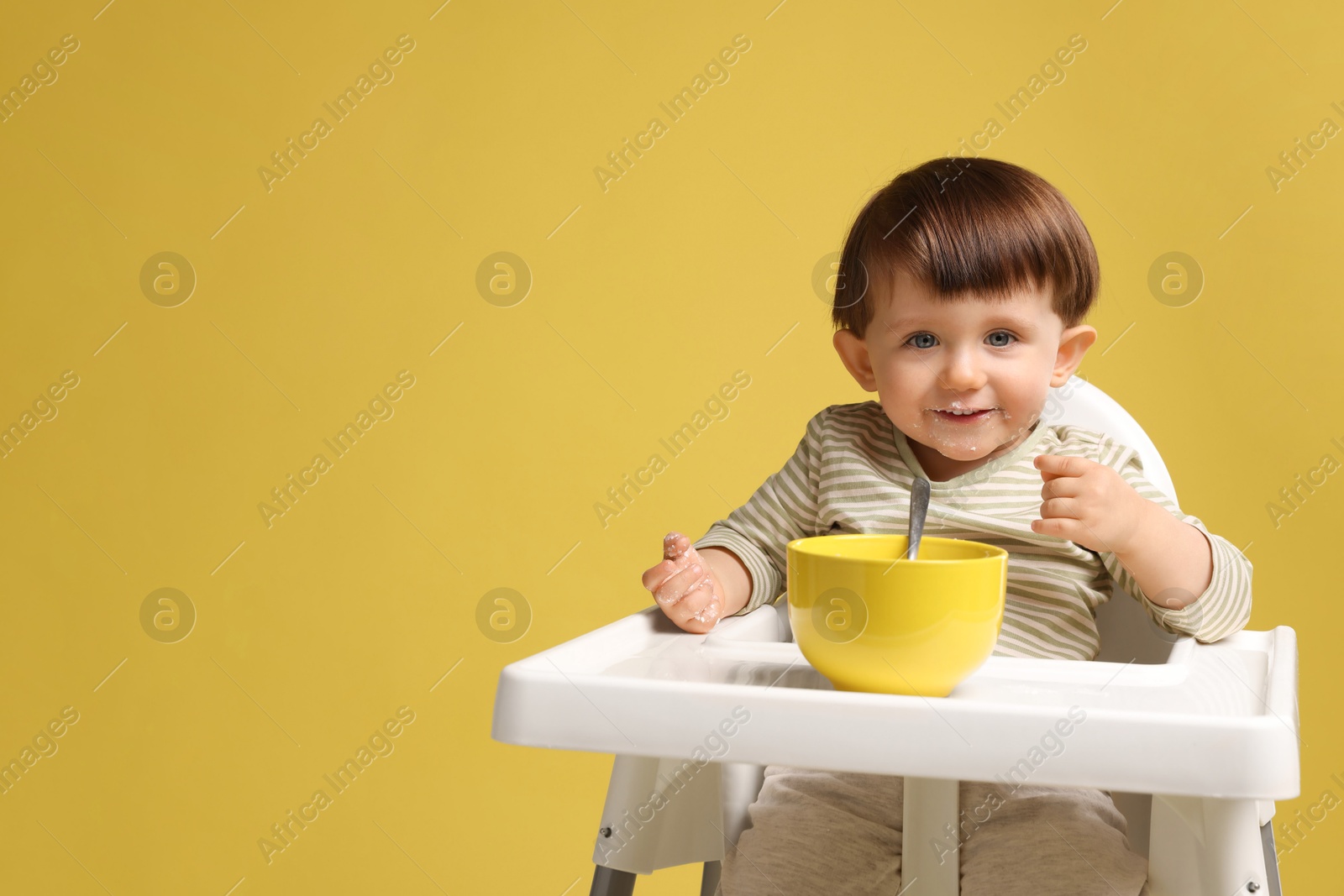 Photo of Cute little kid eating healthy baby food from bowl in high chair on yellow background, space for text