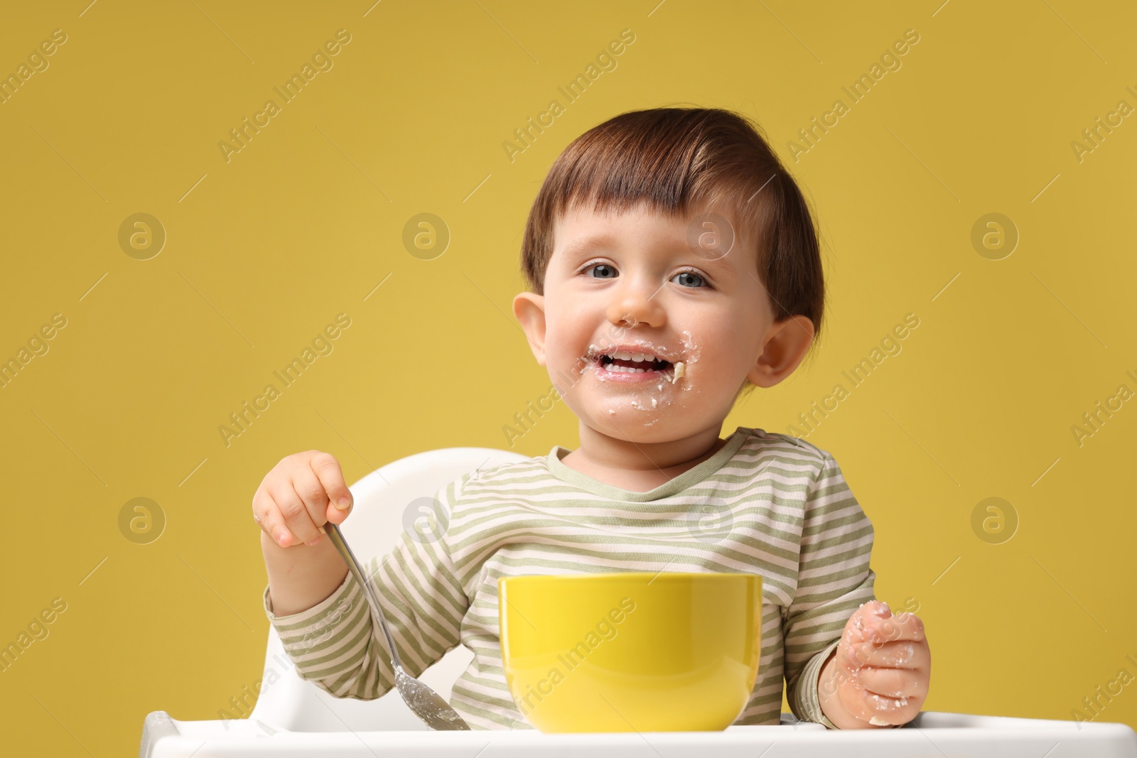 Photo of Cute little kid eating healthy baby food from bowl in high chair on yellow background