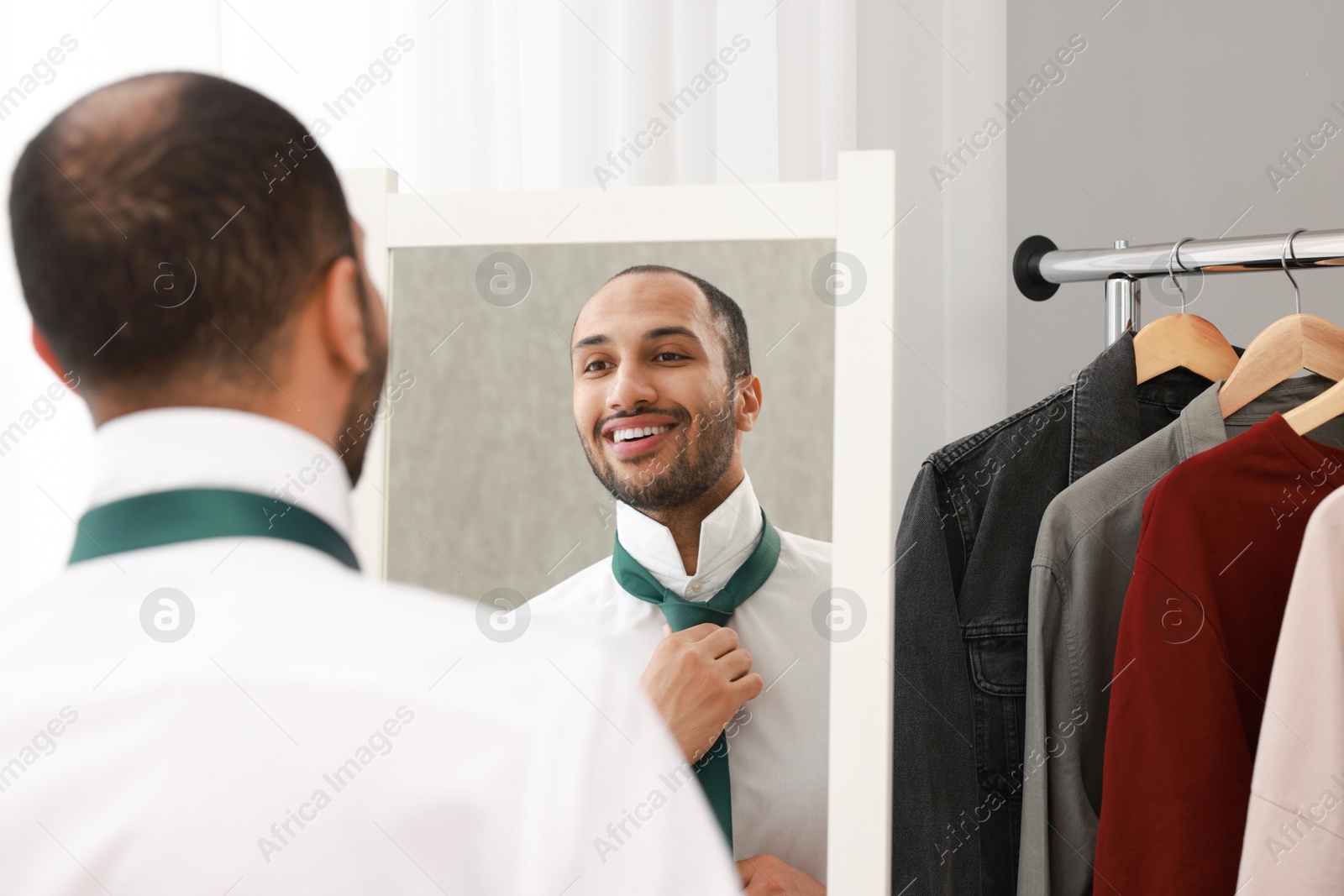 Photo of Smiling man adjusting necktie near mirror at home, back view