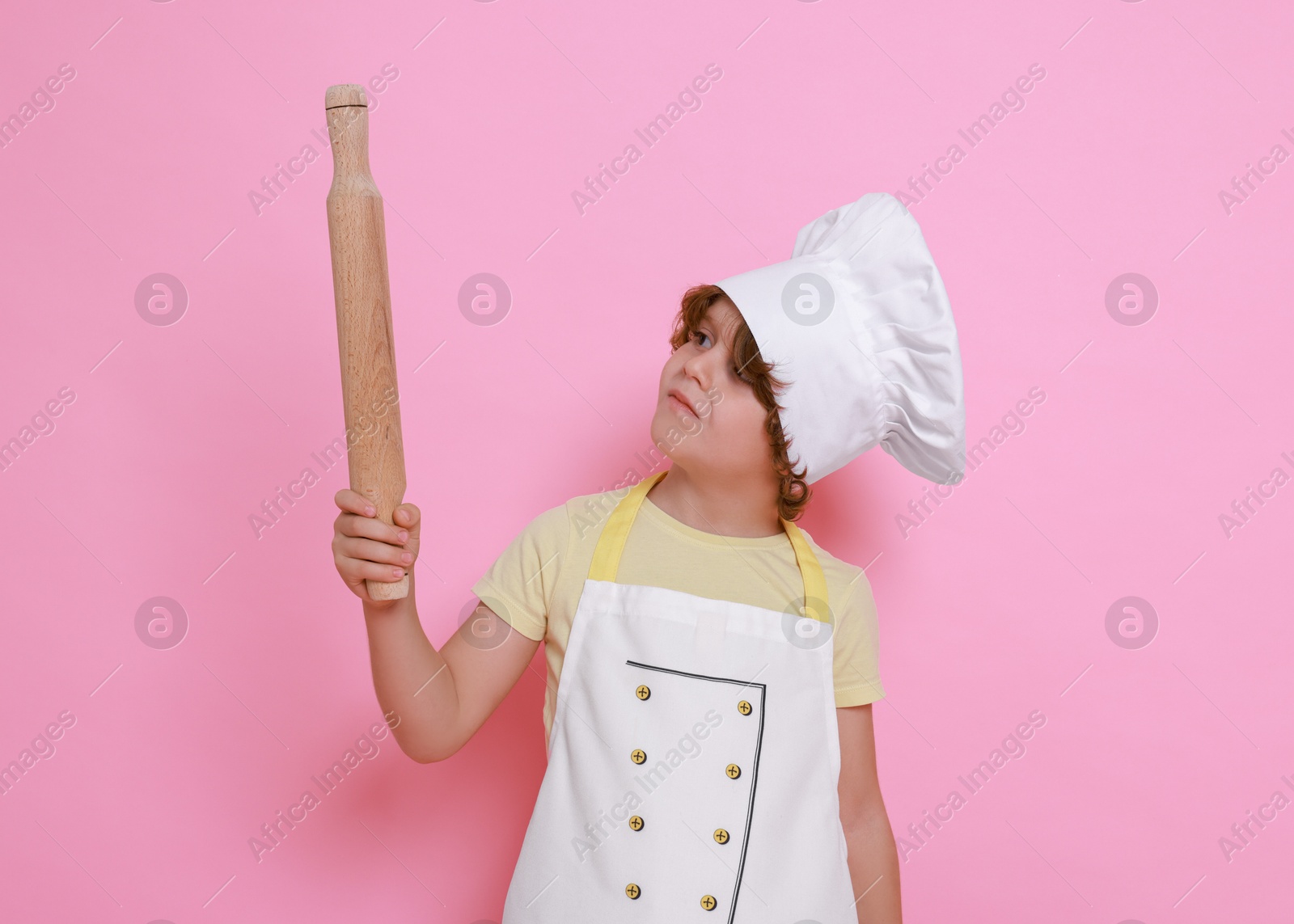 Photo of Little boy with rolling pin pretending to be chef on pink background. Dreaming about future profession