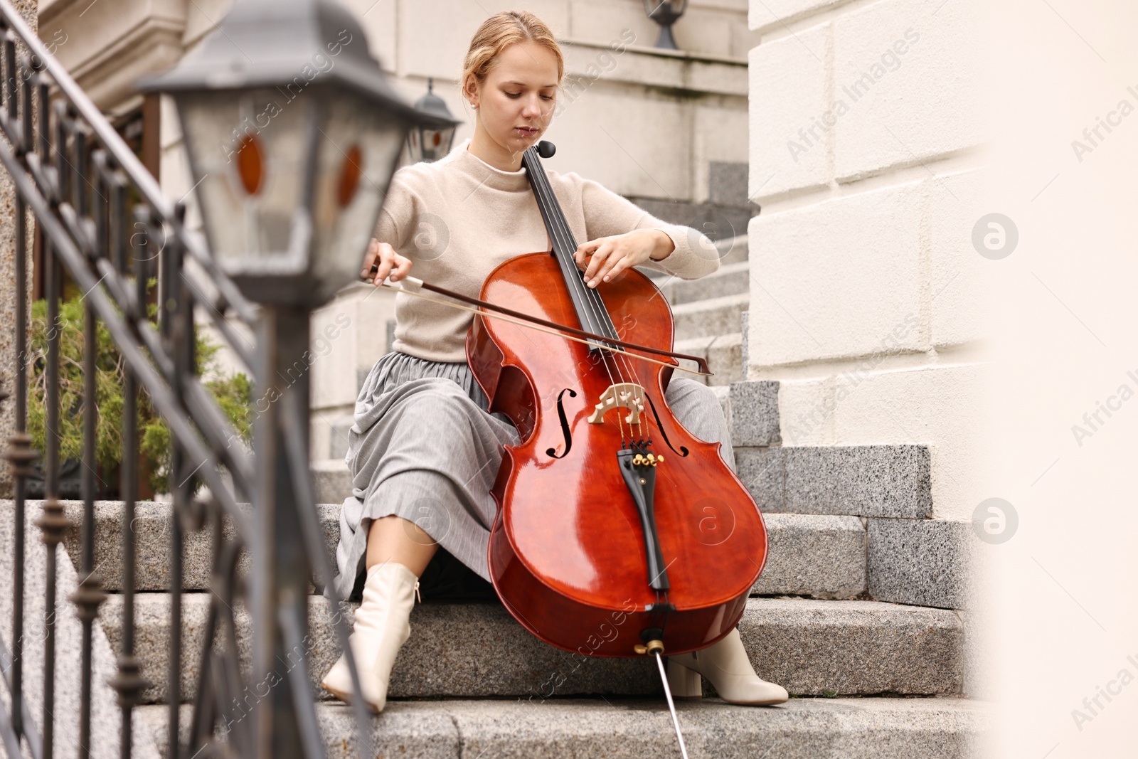 Photo of Beautiful young woman playing cello on stairs outdoors. Classic musical instrument