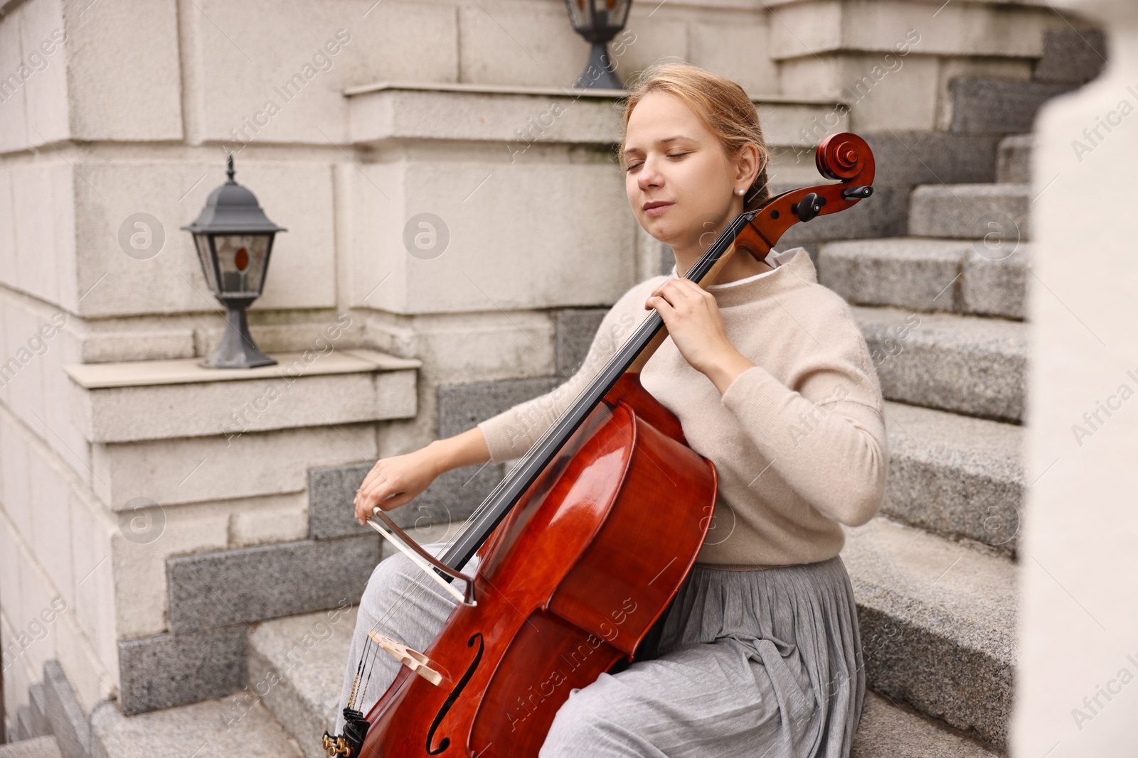 Photo of Beautiful young woman playing cello on stairs outdoors. Classic musical instrument