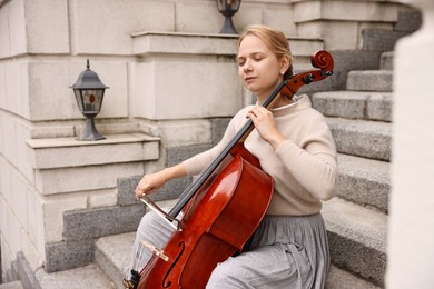 Photo of Beautiful young woman playing cello on stairs outdoors. Classic musical instrument