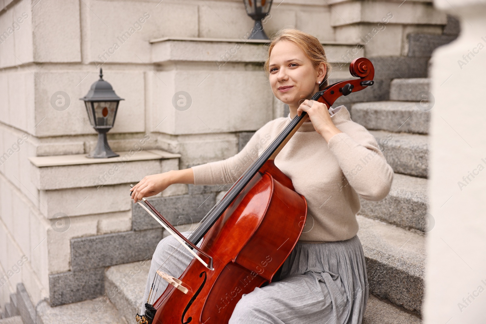 Photo of Beautiful young woman playing cello on stairs outdoors. Classic musical instrument