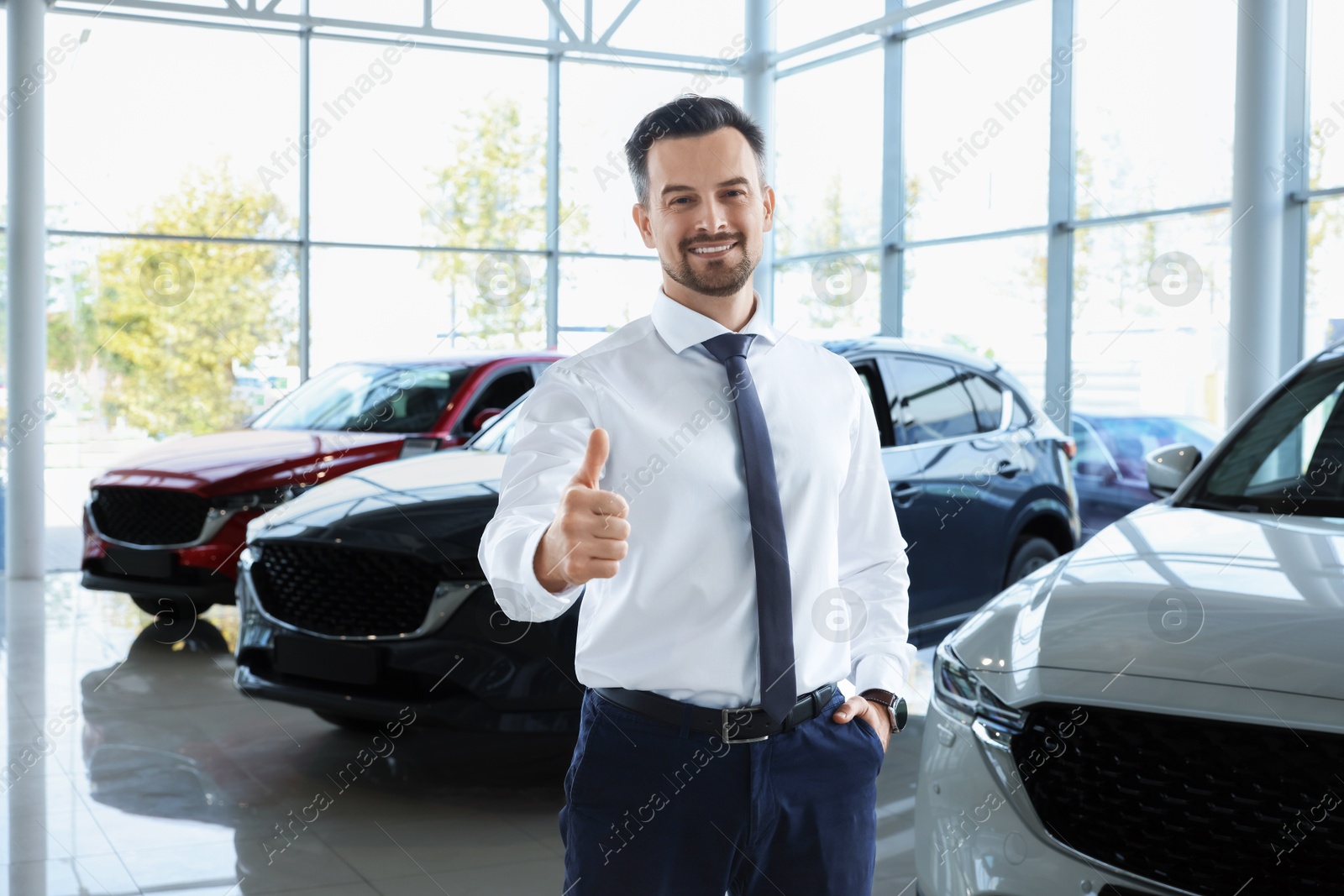 Photo of Happy salesman showing thumbs up near new cars in salon