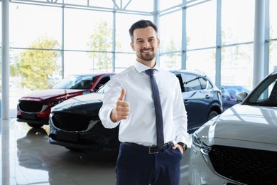 Happy salesman showing thumbs up near new cars in salon