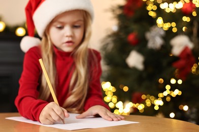 Photo of Little girl writing letter to Santa Claus at table indoors, selective focus. Christmas celebration