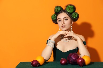 Photo of Young woman with peppers in her hair, fruits and vegetables at green table on orange background, space for text