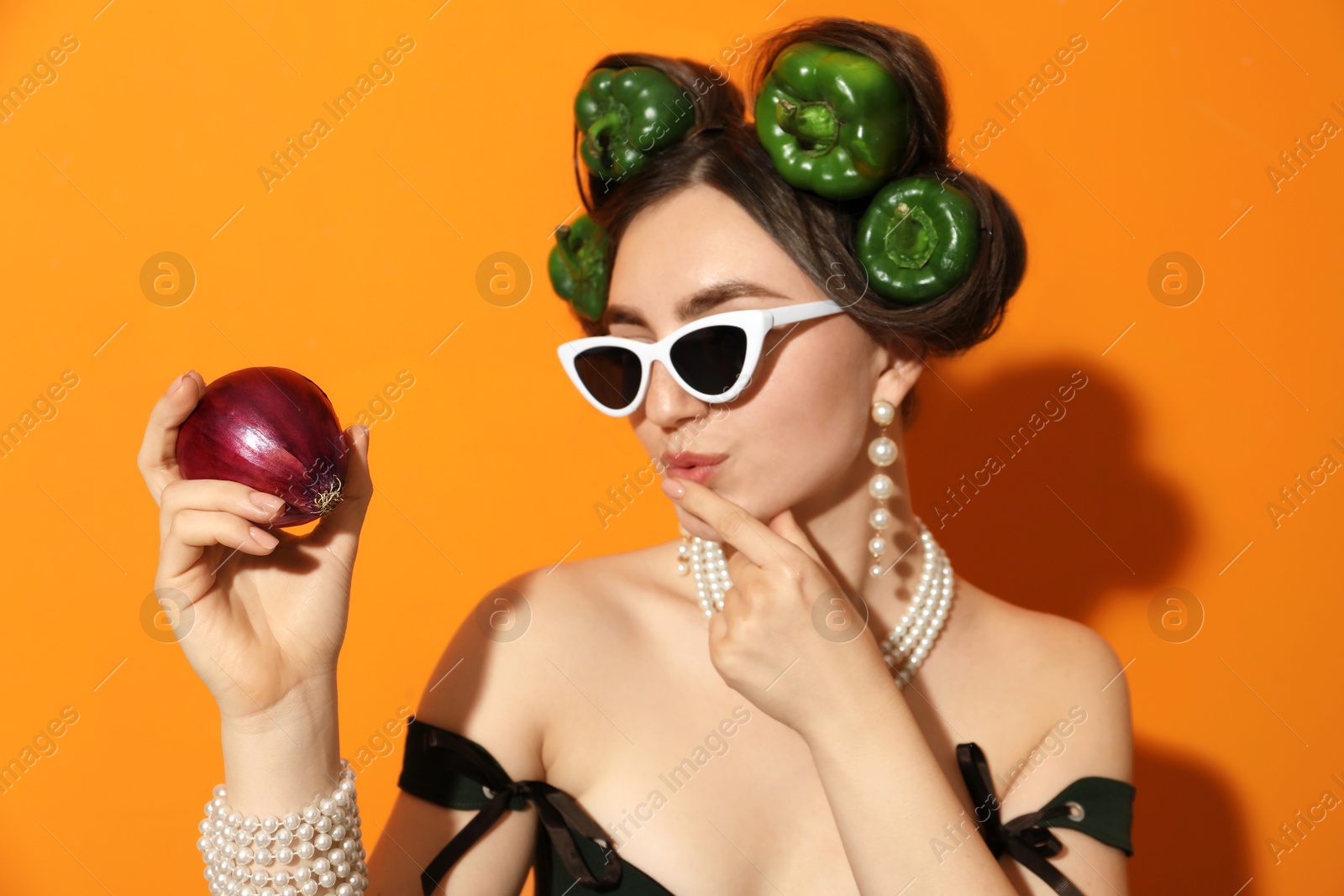 Photo of Young woman with peppers in her hair, sunglasses and onion on orange background