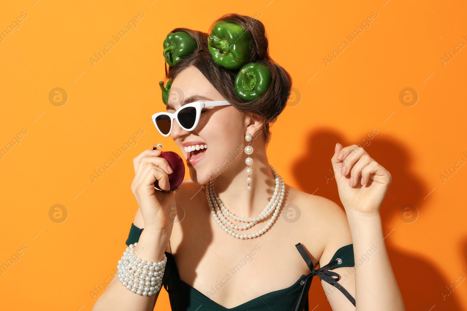 Photo of Young woman with peppers in her hair, sunglasses and onion on orange background