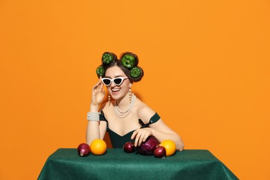 Photo of Young woman with peppers in her hair, sunglasses, fruits and vegetables at green table on orange background