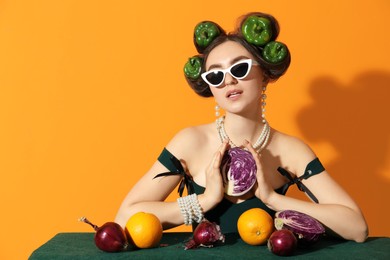 Photo of Young woman with peppers in her hair, sunglasses, fruits and vegetables at green table on orange background, space for text