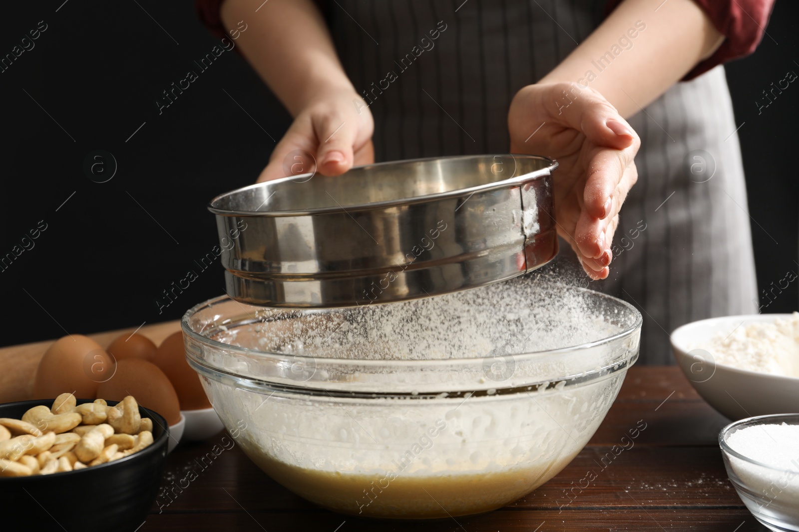 Photo of Making cashew cookies. Woman sieving flour into dough at wooden table, closeup