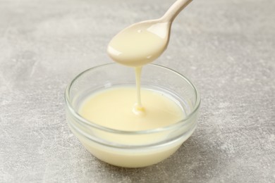 Photo of Condensed milk flowing down from spoon into bowl on gray textured table, closeup