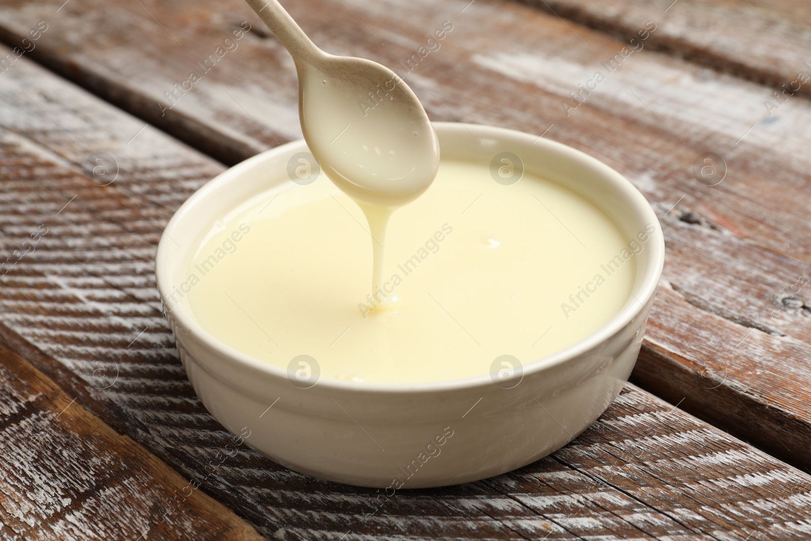 Photo of Condensed milk flowing down from spoon into bowl on wooden table, closeup