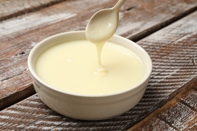 Photo of Condensed milk flowing down from spoon into bowl on wooden table, closeup