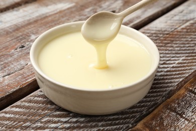 Photo of Condensed milk flowing down from spoon into bowl on wooden table, closeup