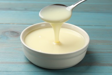 Photo of Condensed milk flowing down from spoon into bowl on light blue wooden table, closeup