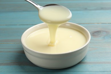 Photo of Condensed milk flowing down from spoon into bowl on light blue wooden table, closeup