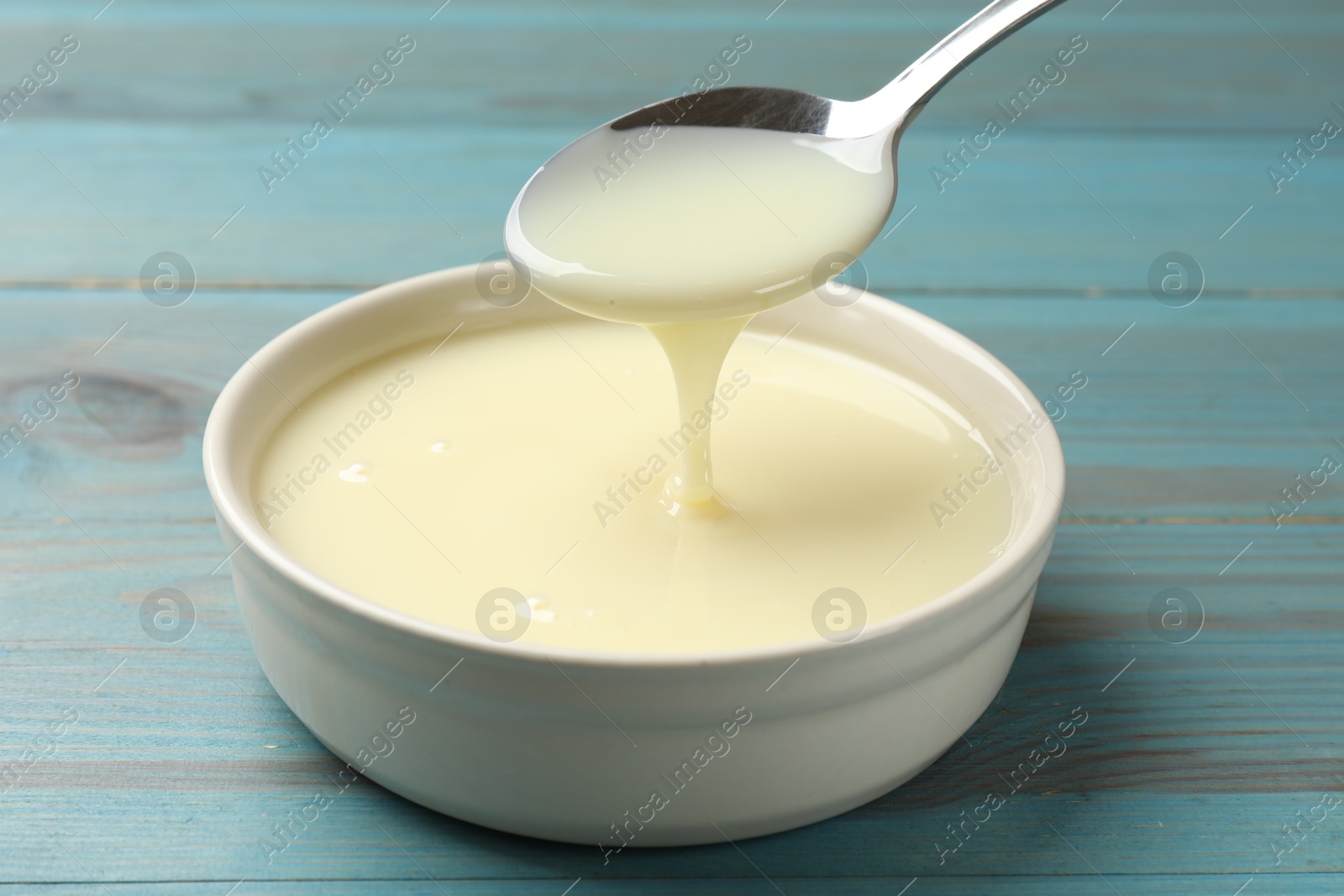 Photo of Condensed milk flowing down from spoon into bowl on light blue wooden table, closeup