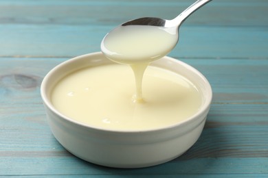 Photo of Condensed milk flowing down from spoon into bowl on light blue wooden table, closeup