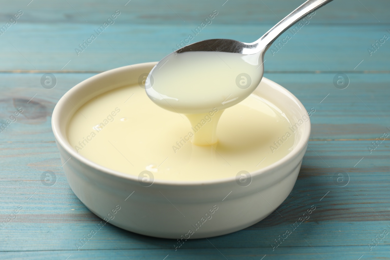 Photo of Condensed milk flowing down from spoon into bowl on light blue wooden table, closeup