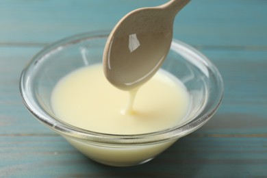 Photo of Condensed milk flowing down from spoon into bowl on light blue wooden table, closeup