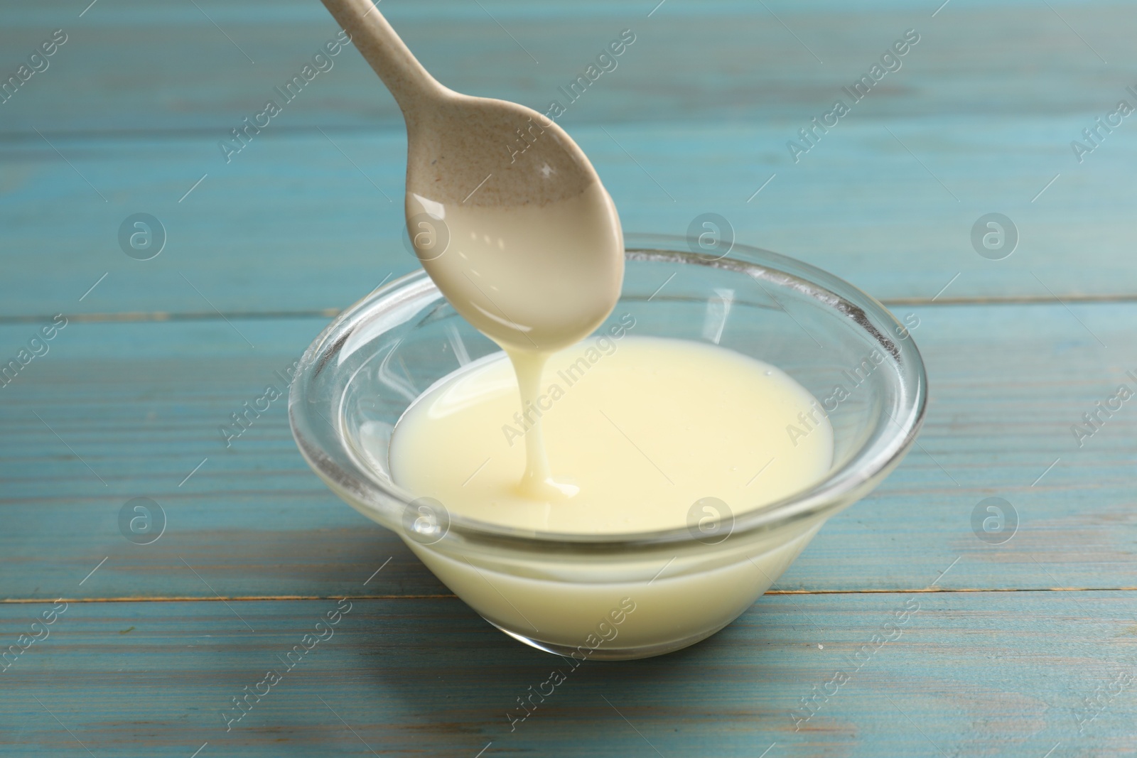 Photo of Condensed milk flowing down from spoon into bowl on light blue wooden table, closeup