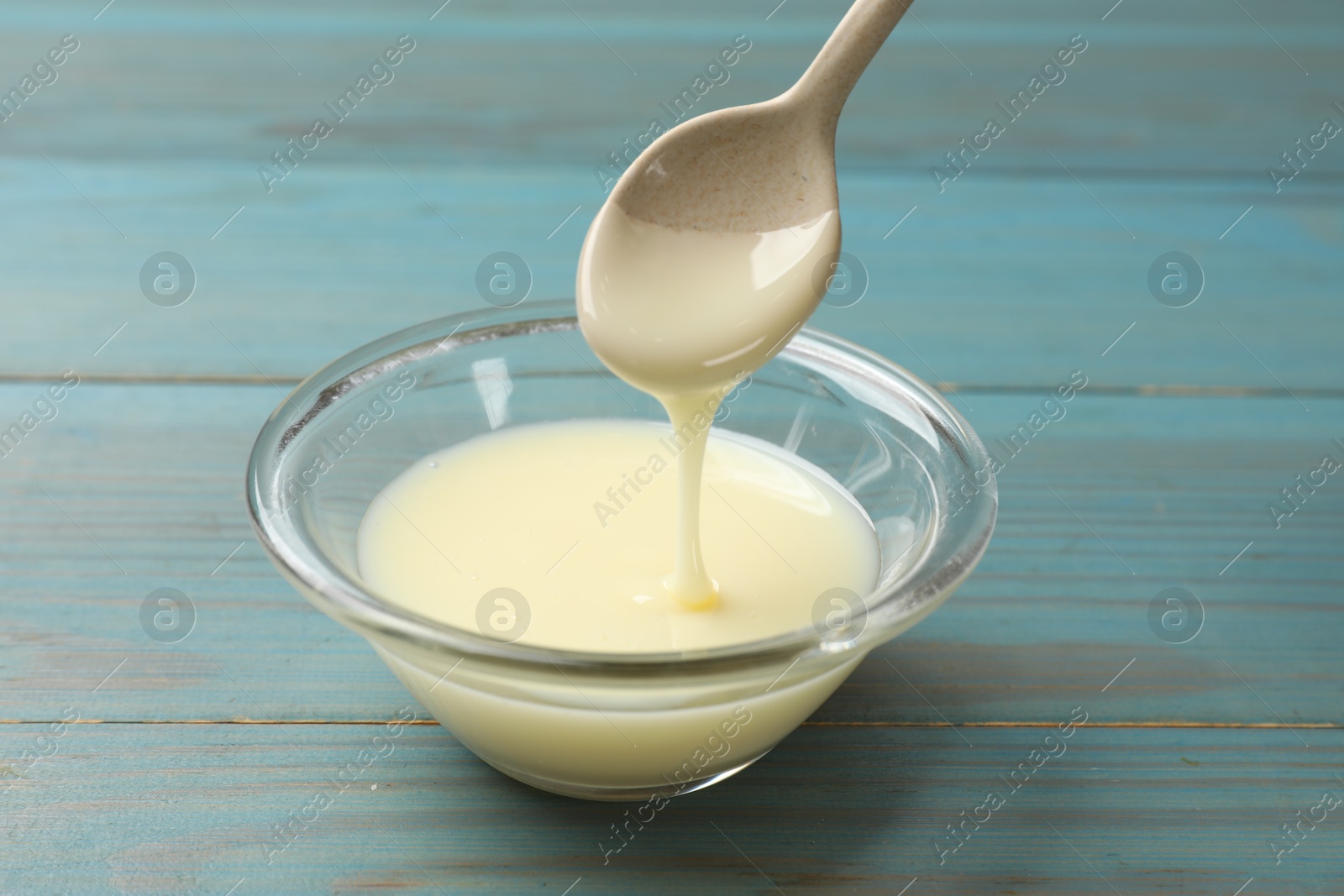 Photo of Condensed milk flowing down from spoon into bowl on light blue wooden table, closeup