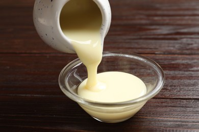 Photo of Pouring delicious condensed milk from jug into bowl on wooden table, closeup