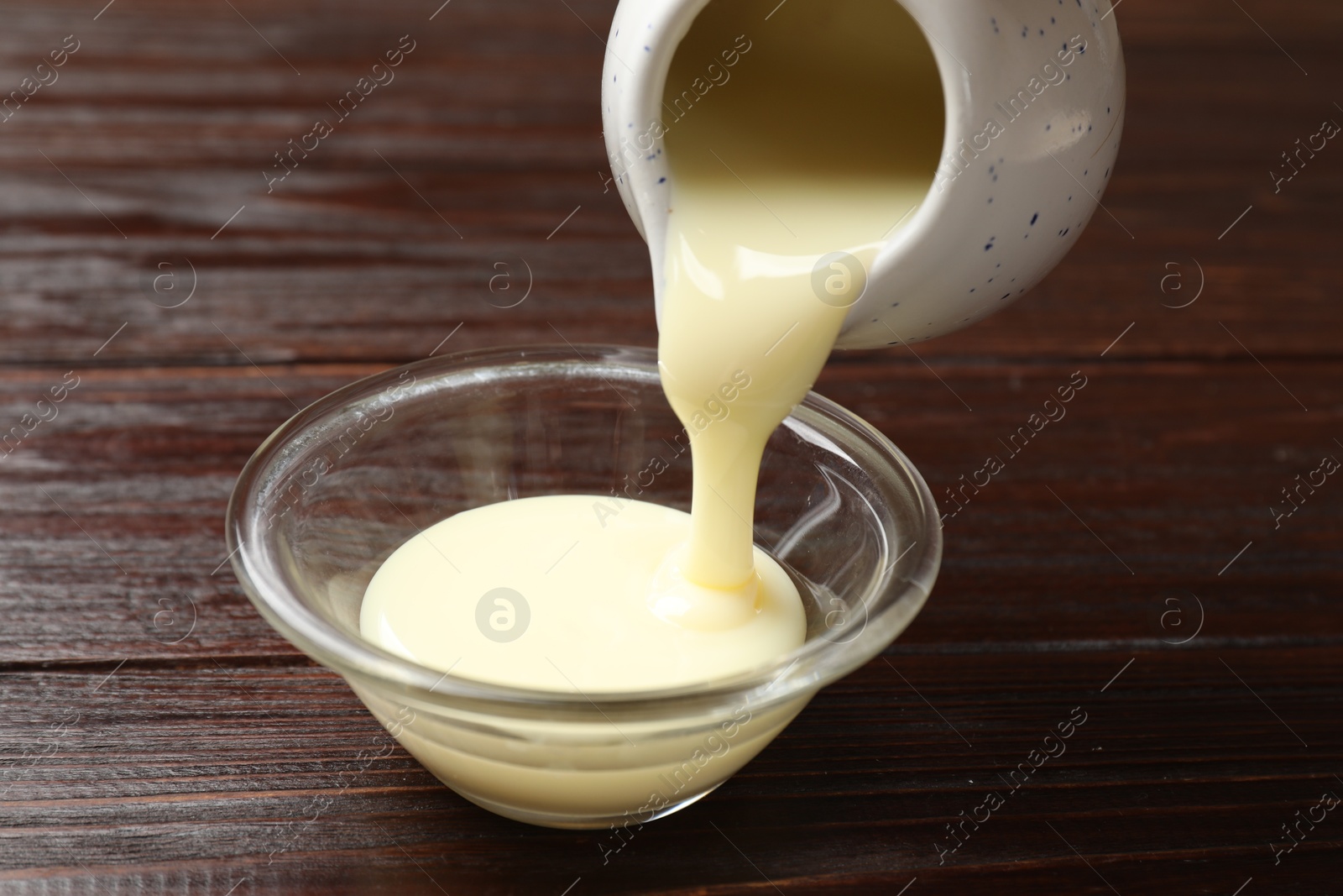 Photo of Pouring delicious condensed milk from jug into bowl on wooden table, closeup