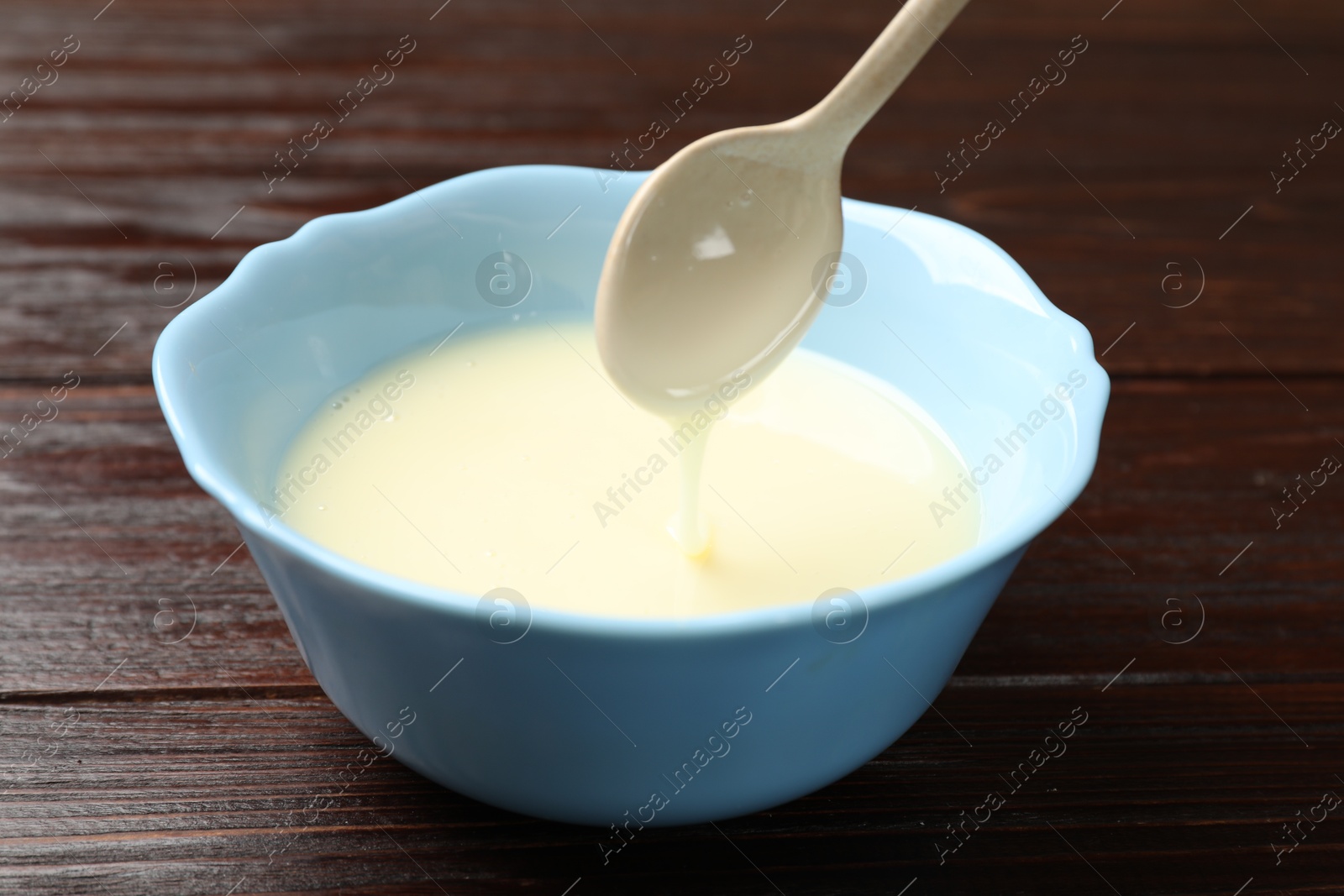 Photo of Condensed milk flowing down from spoon into bowl on wooden table, closeup