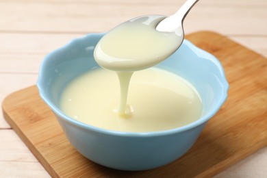 Photo of Condensed milk flowing down from spoon into bowl on wooden table, closeup